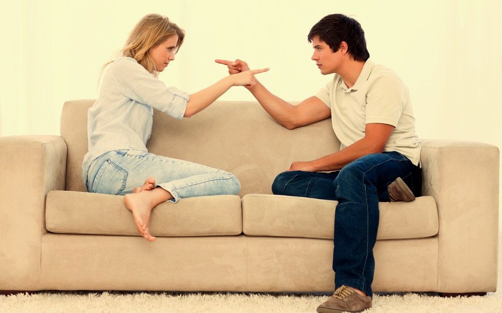 A couple sitting apart on a sofa mid argument, pointing at each other